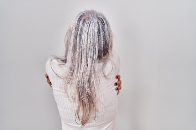 Frau mittleren Alters mit grauem Haar, die vor weißem Hintergrund steht und sich glücklich und positiv von rückständiger Selbstliebe und Selbstfürsorge umarmt