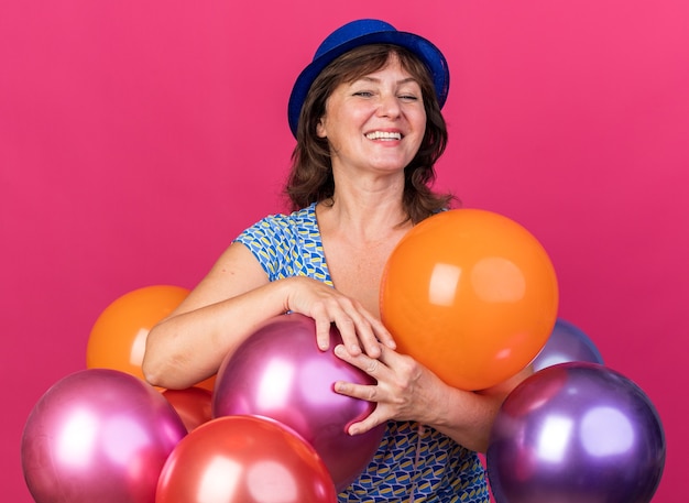 Frau mittleren Alters mit Partyhut, die bunte Luftballons mit glücklichem Gesicht hält, das fröhlich lächelt