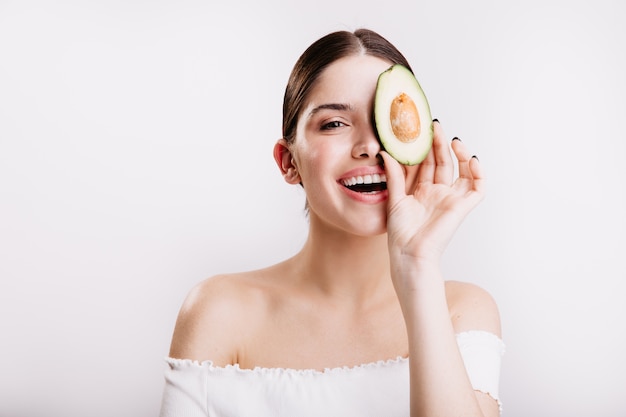 Frau ohne Make-up mit sauberer Haut lächelt und posiert mit Scheibe Avocado für Porträt auf weißer Wand.