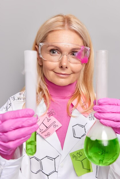 Frau Professor arbeitet an neuen wissenschaftlichen Forschungen vergleicht Flüssigkeit in Kolben und Tube trägt transparente Brillen Gummihandschuhe und weißen Kittel Posen im Labor auf Grau