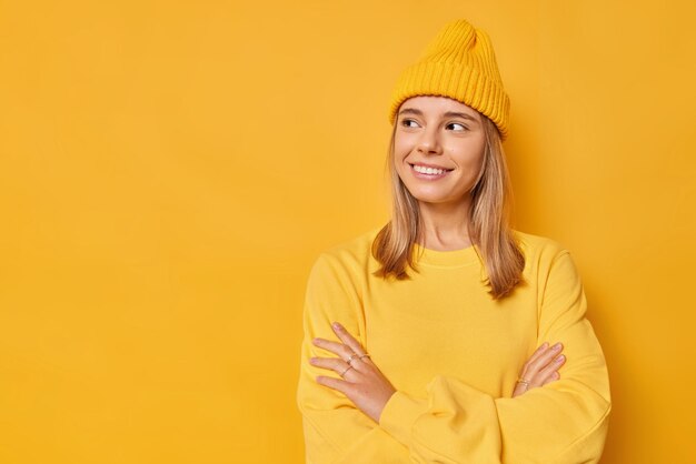 Frau schaut mit frohem Ausdruck weg, hält die Arme verschränkt, hat einen nachdenklichen Look, trägt einen lässigen Pullover und einen Hut, isoliert auf leuchtendem Gelb mit Kopierraum