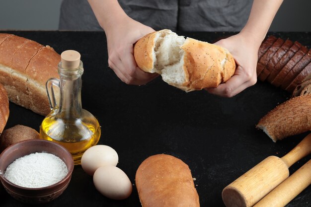 Frau schnitt Brot in zwei Hälften auf dunklem Tisch mit Eiern, Mehlschale und Glas Öl.