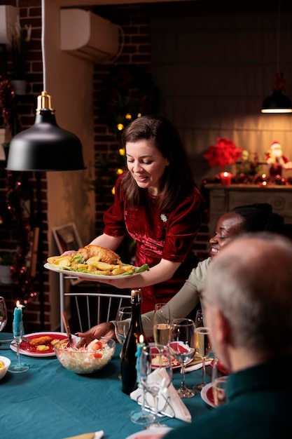 Kostenloses Foto frau serviert festliches essen am tisch und feiert weihnachtsessen mit verschiedenen freunden und familie zu hause. menschen freuen sich, während der weihnachtsfeier hausgemachtes essen zu essen und wein zu trinken.