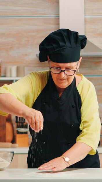 Frau siebt Mehl auf Holztisch in der modernen Küche. Glücklicher älterer Bäcker mit Knochen, der Rohzutaten zum Backen von hausgemachtem Kuchenbestreuen zubereitet, Weizenmehl von Hand sieben