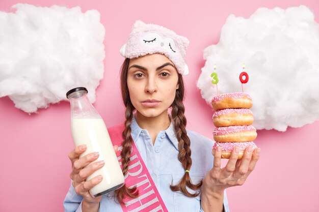 Frau sieht direkt aus hält köstliche Donuts mit Kerzen feiert 30-jähriges Jubiläum trägt Schlafmaske und wird eine festliche Schlafmaske trinkt frische Milch isoliert auf rosa Wand