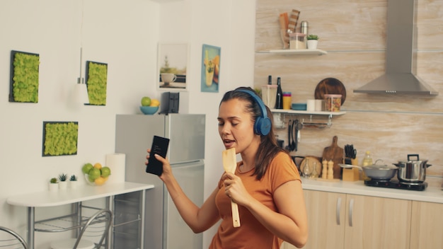 Frau singt morgens mit Holzlöffel. Energiegeladene, positive, fröhliche, lustige und süße Hausfrau, die alleine im Haus tanzt. Unterhaltung und Freizeit allein zu Hause