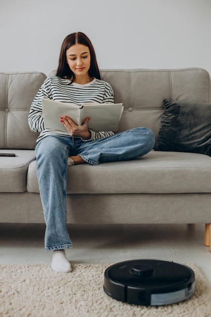 Frau sitzt auf dem Sofa und liest ein Buch, während der Roboterstaubsauger den Raum aufräumt