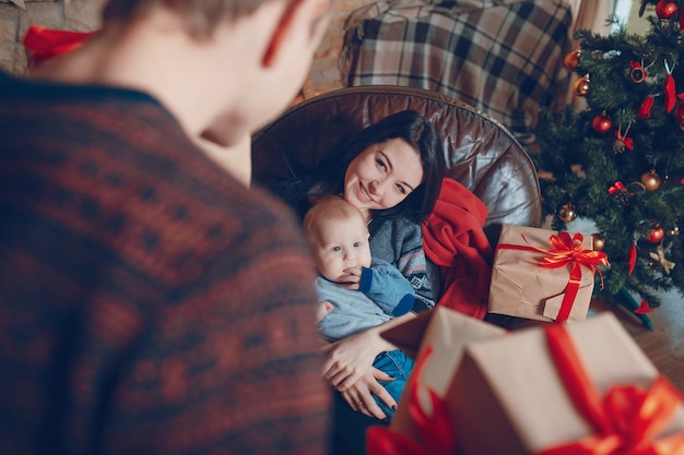 Frau sitzt auf einem Sofa mit einem Baby auf dem Arm und mit einem Berg von braunen Geschenke mit roter Schleife