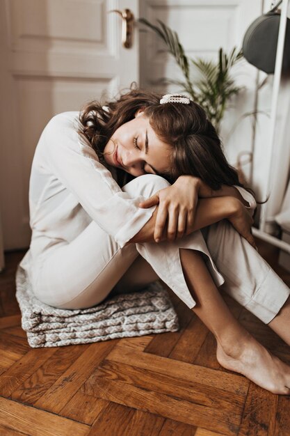 Frau sitzt auf Holzboden und schläft Hübsches junges Mädchen in beigem Hemd und Hose, das sich in einer gemütlichen weißen Wohnung ausruht
