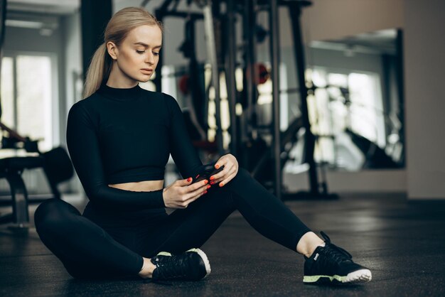 Frau sitzt im Fitnessstudio auf dem Boden und benutzt Handy
