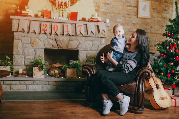 Frau sitzt in einem Sessel mit ihrem Baby und einer Gitarre neben und einem Kamin Hintergrund