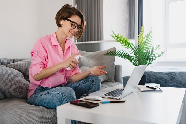 Frau sprüht Desinfektionsmittel antiseptisch auf die Hände am Arbeitsplatz zu Hause, die online am Laptop arbeitet