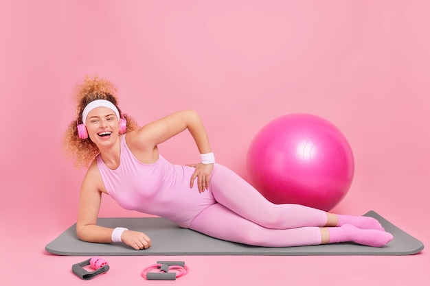 Frau steht in Plankenpose auf Fitnessmatte in Activewear gekleidet hört Musik über Kopfhörer benutzt Sportgeräte