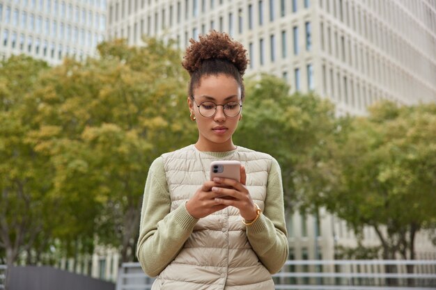 Frau surft im Internet auf einem modernen Smartphone beeilt sich zum Treffen trägt einen runden Brillenpullover und eine Weste posiert im Freien gegen moderne Gebäude und Bäume