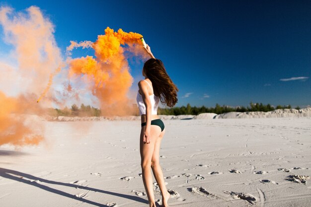 Frau tanzt mit orange Rauch am weißen Strand unter blauem Himmel