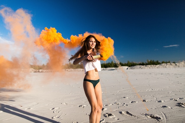 Frau tanzt mit orange Rauch am weißen Strand unter blauem Himmel