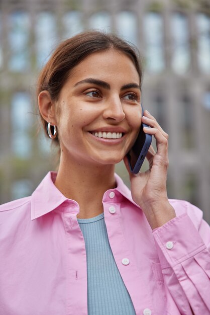 Frau telefoniert mit modernem Smartphone-Lächeln trägt angenehm rosa Hemdposen auf der Straße und ist gut gelaunt. Menschen Technologie Lifestyle-Konzept.