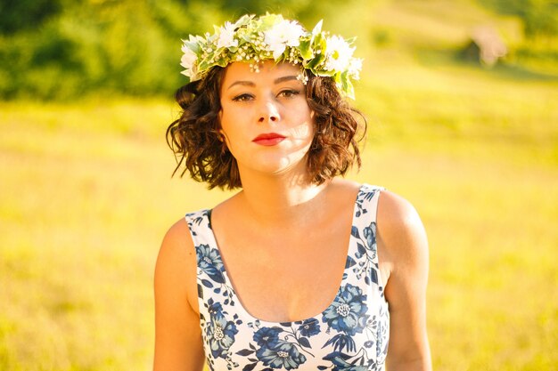 Frau trägt ein Blumenkleid mit einem Blumenkranz auf dem Kopf und posiert auf einem Feld
