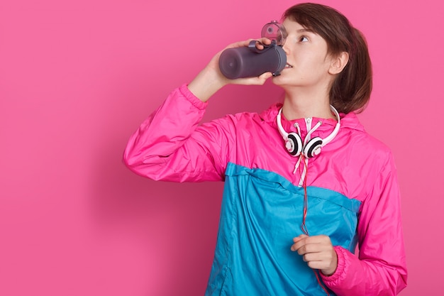 Frau trägt in Blau und Rose Sportswear Trinkwasser aus der Flasche, Modell posiert isoliert auf rosig. Junge weibliche Fitnesstrainerin oder Personal Trainerin im Studio.