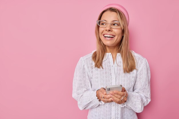 Frau trägt transparente Brille Stirnband weiße Bluse hält moderne Handy-Chats online mit Freunden überprüft die E-Mail-Box sieht glücklich weg isolierte rosa Wand