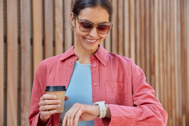 Frau überprüft die Zeit auf der Uhr hält Kaffee zum Mitnehmen genießt Koffeingetränk trägt eine Sonnenbrille rosa Hemd wird sich mit einem Freund treffen lächelt angenehm
