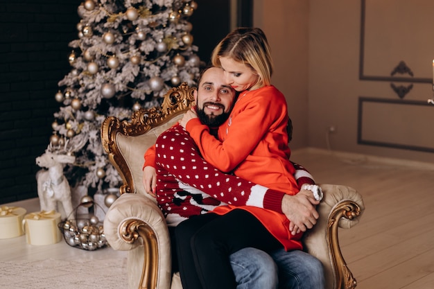 Frau umarmt ihren Mann, der zart im weichen großen Stuhl vor einem Weihnachtsbaum sitzt