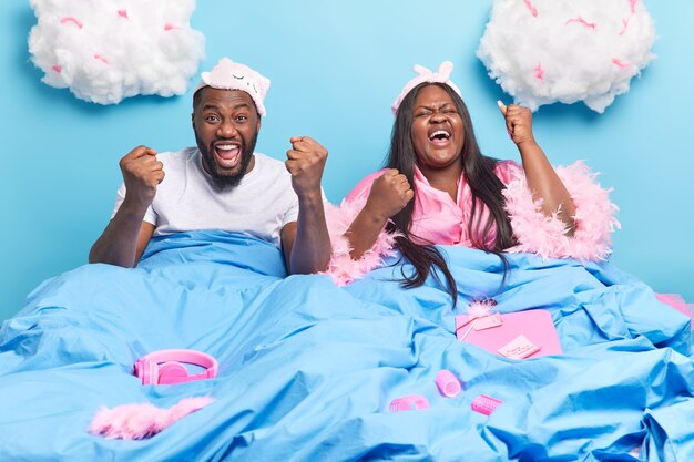 Frau und Mann ballen die Fäuste, freuen sich über das Wochenende und die Freizeit posieren im Bett unter einer weichen Decke, die in häuslicher Kleidung isoliert auf Blau gekleidet ist