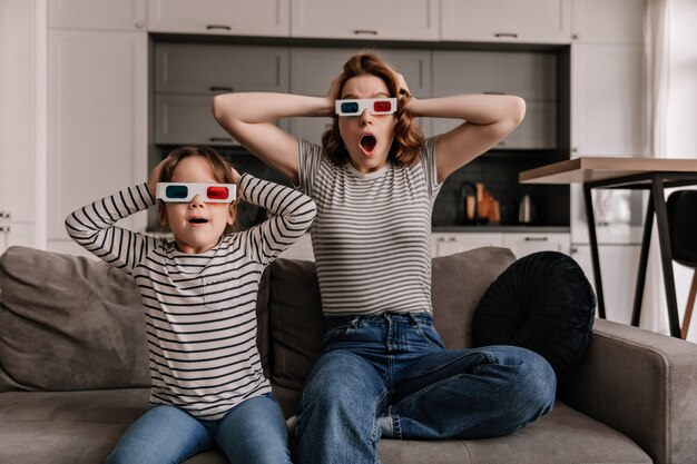 Frau und weibliches Kind sind schockiert, wenn sie einen Film in 3D ansehen und auf dem Sofa in der Wohnung sitzen.