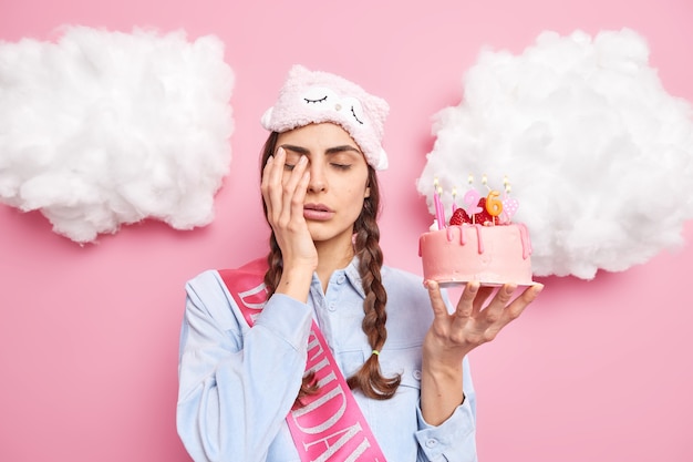 Frau wacht sehr früh an ihrem geburtstag auf nachteile gesicht mit hand schließt augen hält festlichen kuchen trägt schlafmaske auf der stirn hat zwei gekämmte zöpfe isoliert auf rosa