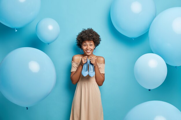 Frau wählt Outfit für Date trägt braunes Cocktailkleid hält blaue hochhackige Schuhe bereitet sich auf Partyposen vor