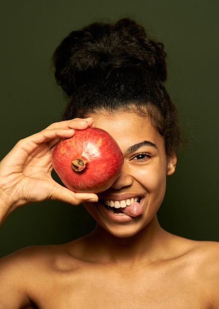 Frau zeigt ihre Zunge, hält Granatapfelfrüchte auf ihrem Auge und posiert isoliert auf Grün