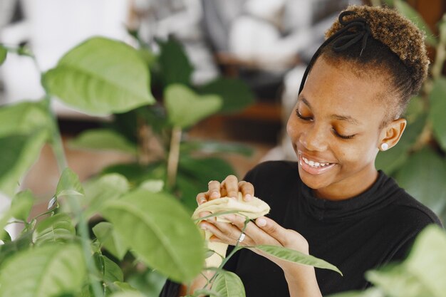 Frau zu Hause. Mädchen in einem schwarzen Pullover. Afrikanerin benutzt den Lappen. Person mit Blumentopf.