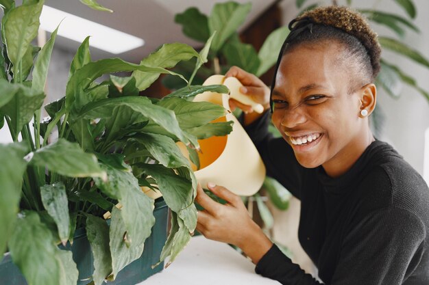 Frau zu Hause. Mädchen in einem schwarzen Pullover. Afrikanische Frau, die die Pflanze gießt. Person mit Blumentopf.