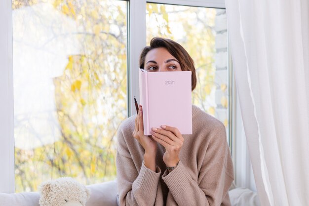 Frau zu Hause mit Notizbuch sitzen auf der Fensterbank in bequemem Pullover und warmen Wollsocken, kalter Herbst außerhalb des Fensters