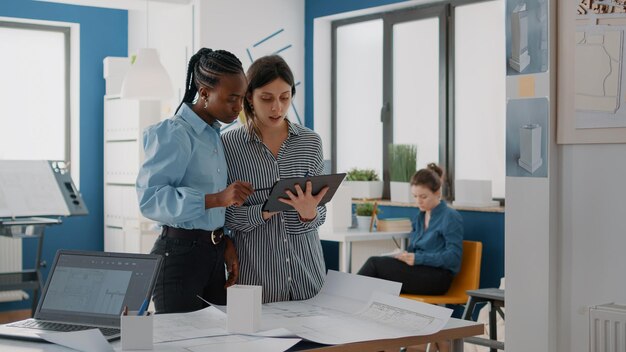 Frauen, die sich ein digitales Tablet und Blaupausen ansehen, um das Gebäudelayout für den Bau zu planen. Kollegen arbeiten zusammen, um Struktur und architektonische Entwicklung zu entwerfen. Architekten bei der Arbeit