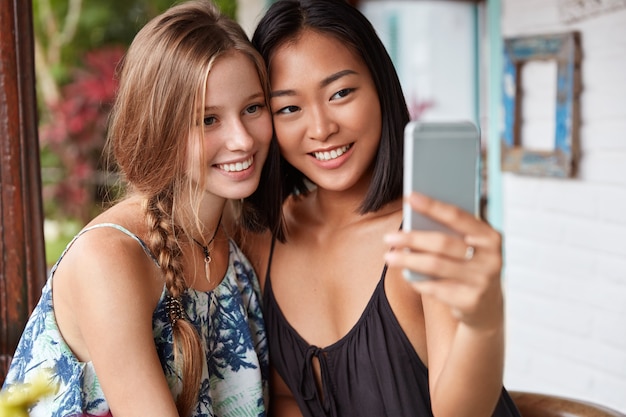 Frauen gemischter Rassen haben eine wahrheitsgemäße Freundschaft und posieren für Selfies in einem modernen Café. Multiethnische junge Frauen benutzen Handy, um Foto zu machen