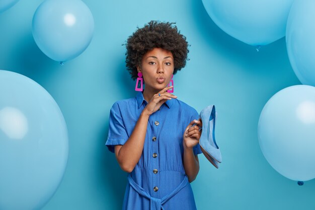 Frauen, Mode, Stil, Partykonzept. Elegante ernsthafte Frau im blauen Kleid, hält hochhackige Schuhe, Kleider für Themenparty, bereit zum Ausgehen, posiert gegen blaue Wand mit Heliumballons