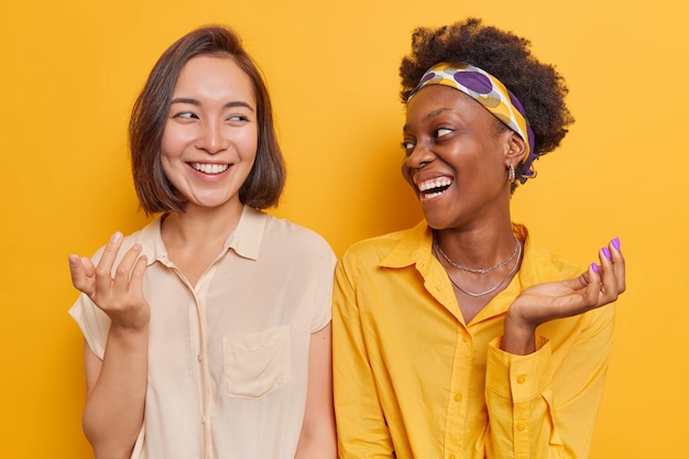 Kostenloses Foto frauen sehen sich gerne in stilvollen kleidern an, lächeln im großen und ganzen, haben spaß, halten die hände erhoben, stehen schulter an schulter isoliert auf gelbem studio