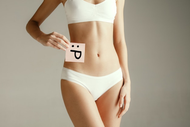 Frauengesundheit. Weibliches Modell, das Karte mit Emoji nahe Magen hält. Junges erwachsenes Mädchen mit Papier für Zeichen oder Symbol lokalisiert auf grauem Studiohintergrund. Körperteil ausschneiden. Medizinisches Problem und Lösung.