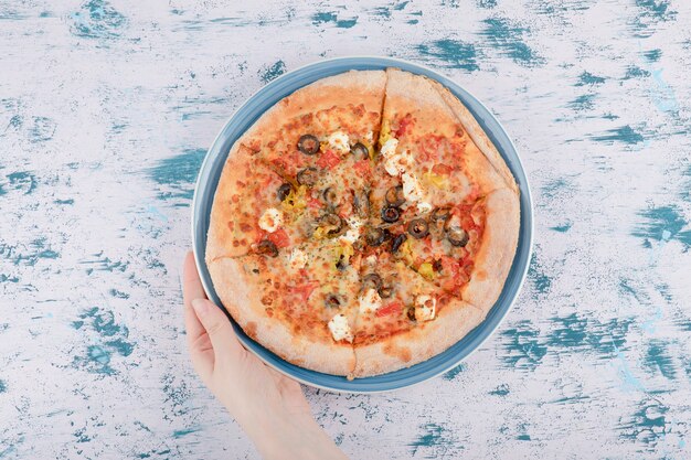 Frauenhand, die einen blauen Teller mit heißer Pizza auf einem Marmorhintergrund d hält.