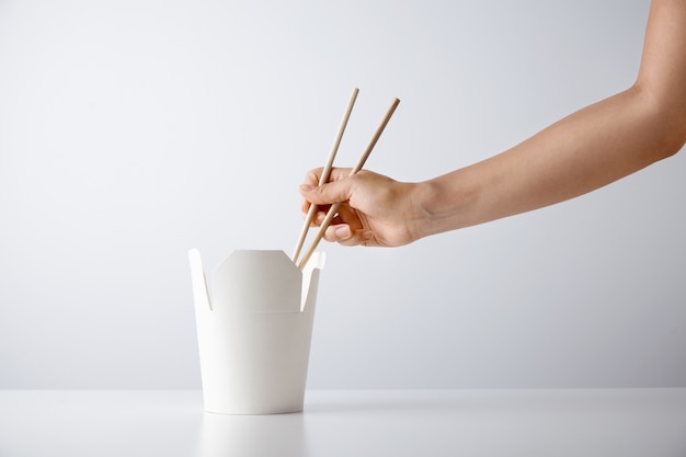 Frauenhand verwendet Essstäbchen, um leckere Nudeln von der leeren Schachtel zum Mitnehmen aufzunehmen, die auf weißer Einzelhandelssetpräsentation isoliert wird