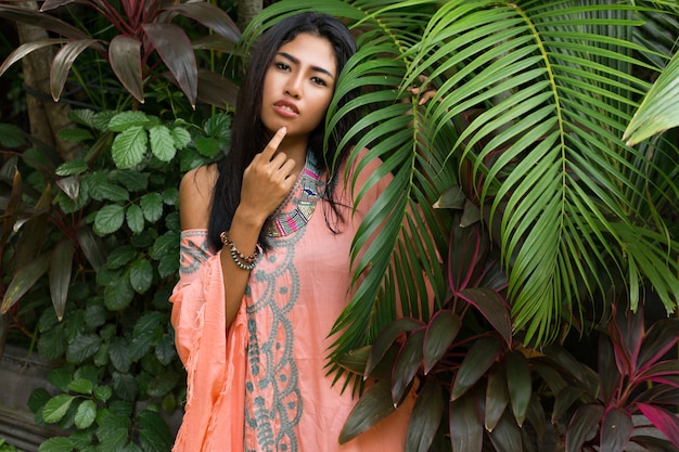 Frauenmodell im Boho-Kleid mit grünen Palmblättern. Schöne asiatische Frau in den modischen Sommerkleidern und -zubehör, die im tropischen Naturporträt aufwerfen.