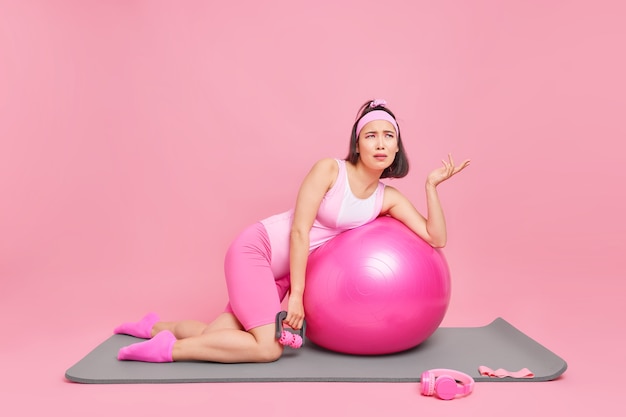 Frauenübungen mit Sportgeräten und Fitnessball hebt die Handfläche hat unzufrieden müde Ausdrucksposen auf Matte über rosa Wand. Heimischer Sport während der Quarantäne