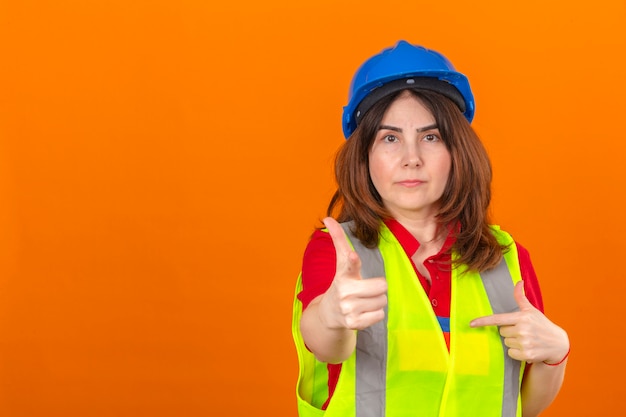 Frauingenieur, der Bauweste und Sicherheitshelm trägt, zeigt mit dem Finger zur Kamera, die ernsthaft über isolierte orange Wand schaut