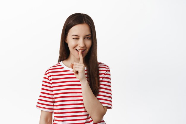 Freche junge Frau zwinkert und schweigt, macht mit dem Finger in der Nähe der Lippen eine shh-Geste, während sie lächelt, hat Überraschung, deutet auf etw hin und steht in rotem T-Shirt vor weißem Hintergrund