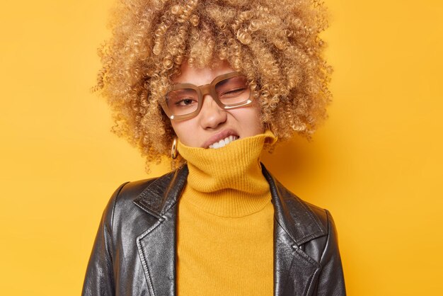Freche lockige Frau zwinkert die Augen bedeckt den Mund mit dem Kragen des Pullovers sieht selbstbewusst in die Kamera, gekleidet in eine stilvolle Lederjacke, einzeln auf leuchtend gelbem Hintergrund. Menschen- und Stilkonzept