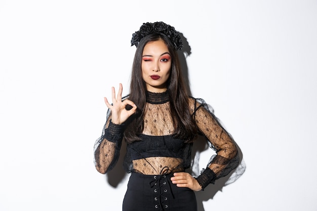 Freche schöne asiatische Frau im Hexenkostüm zeigt okay Geste, alles unter Kontrolle. Zuversichtlich Mädchen in Halloween Gothic Kleid mögen oder genehmigen etwas, stehenden weißen Hintergrund.