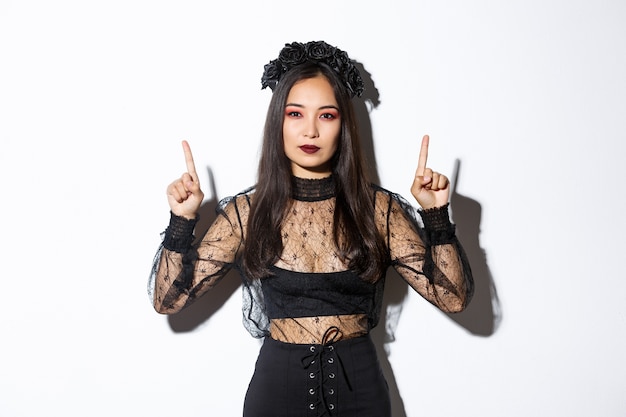 Freche schöne asiatische Frau im schwarzen gotischen Kleid, Hexenkostüm für Halloween tragend und Finger nach oben zeigend