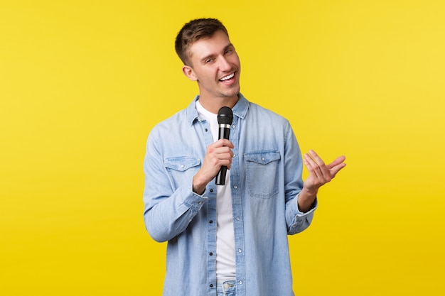 Frecher, gutaussehender blonder Kerl in Freizeitkleidung führt Rede, Stand-up-Show vor dem Publikum, singt ein Lied und lächelt frech, steht auf gelbem Hintergrund, genießt Karaoke-Nacht und hält Mikrofon.