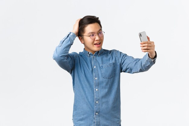 Frecher selbstbewusster asiatischer Mann in Brille und Hosenträger, der sich frech fühlt, Selfie macht, Haare mit der Hand bürstet, für Fotos posiert, Filter-App auf dem Handy verwendet, Blogger im Internet posten.